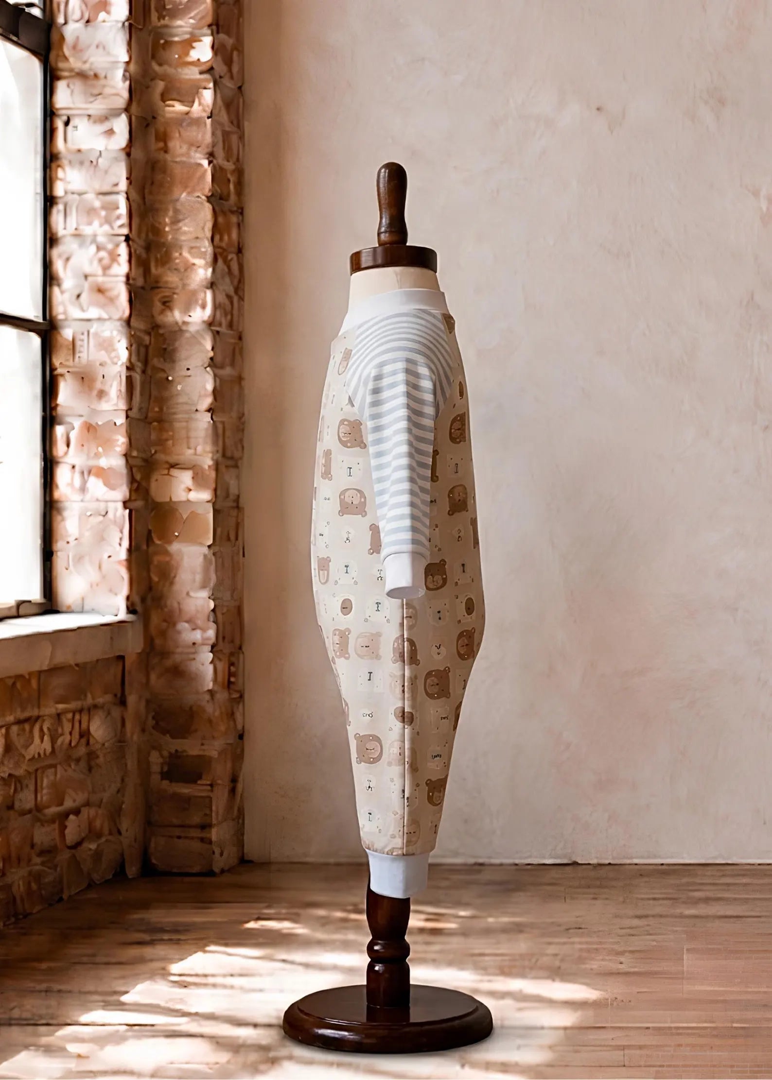 A cream-colored knit sweater with a brown teddy bear pattern and white striped cuffs, displayed on a wooden dress form mannequin in a rustic room with a brick column and textured plaster wall.