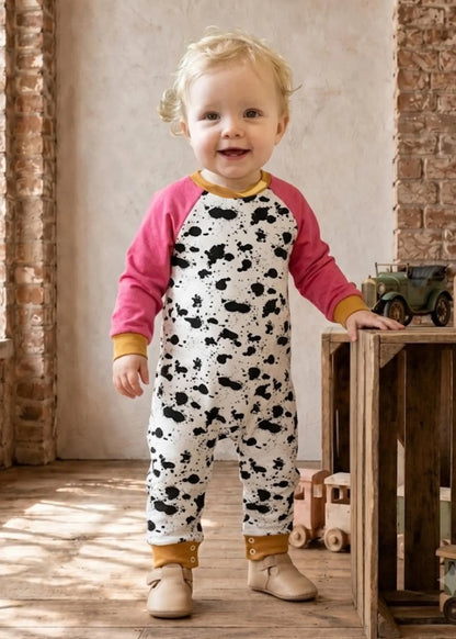 A toddler stands indoors wearing a one-piece jumpsuit with a cow-print body, pink raglan sleeves, and mustard yellow trim. The child is posed beside a wooden crate in a rustic room with exposed brick and wood flooring.