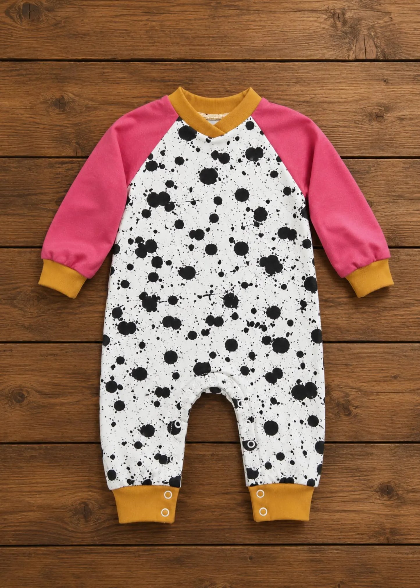 A baby long-sleeve one-piece jumpsuit laid flat on a rustic wooden plank background. The jumpsuit features a white body with a black splatter print, bright pink raglan sleeves, and mustard yellow cuffs and collar.