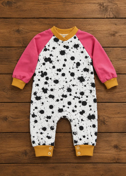 A baby long-sleeve one-piece jumpsuit laid flat on a rustic wooden plank background. The jumpsuit features a white body with a black splatter print, bright pink raglan sleeves, and mustard yellow cuffs and collar.