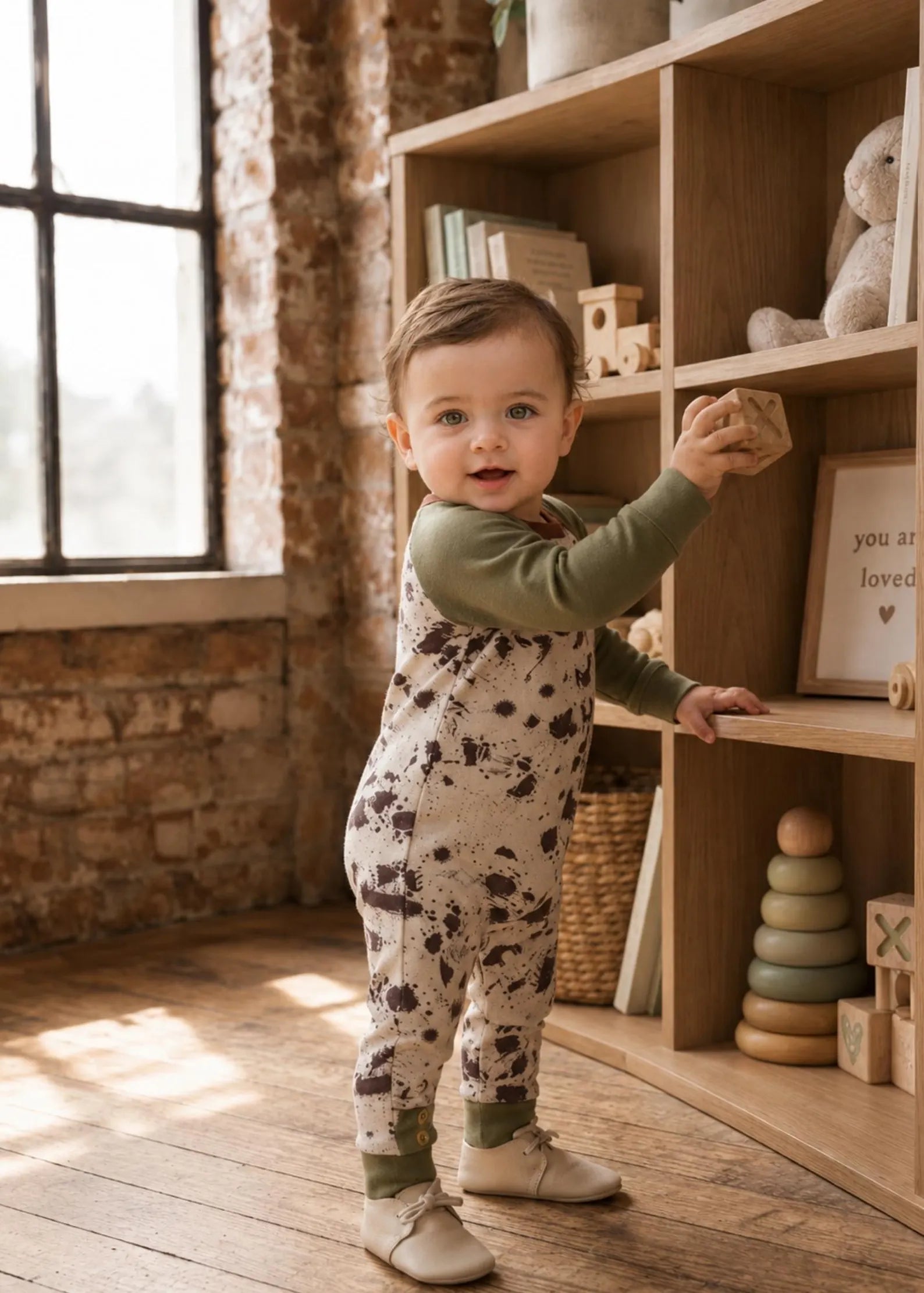 A toddler stands in a sunlit nursery or playroom wearing an olive green long-sleeve jumpsuit with a beige and brown speckled print. The child is posed next to a wooden bookshelf filled with toys and books.