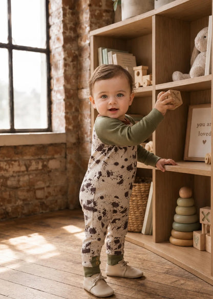 A toddler stands in a sunlit nursery or playroom wearing an olive green long-sleeve jumpsuit with a beige and brown speckled print. The child is posed next to a wooden bookshelf filled with toys and books.