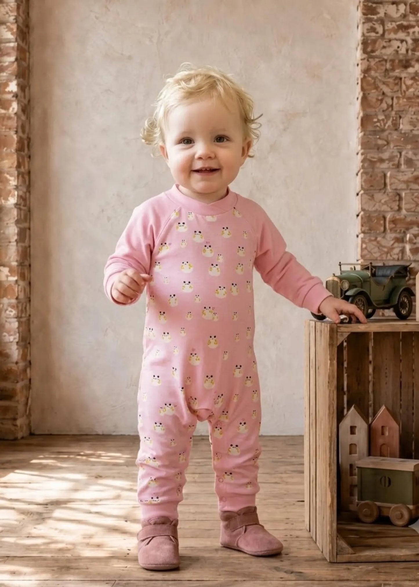 A smiling toddler stands indoors wearing a pink long-sleeve footless one-piece jumpsuit covered in small owl illustrations. The child is posed beside a wooden crate with toy vehicles against a rustic wall and wood floor.