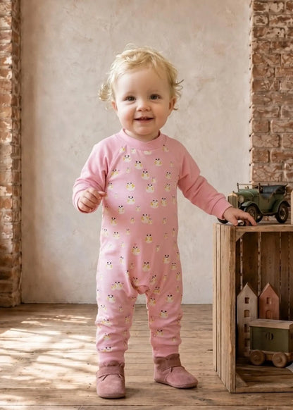 A smiling toddler stands indoors wearing a pink long-sleeve footless one-piece jumpsuit covered in small owl illustrations. The child is posed beside a wooden crate with toy vehicles against a rustic wall and wood floor.