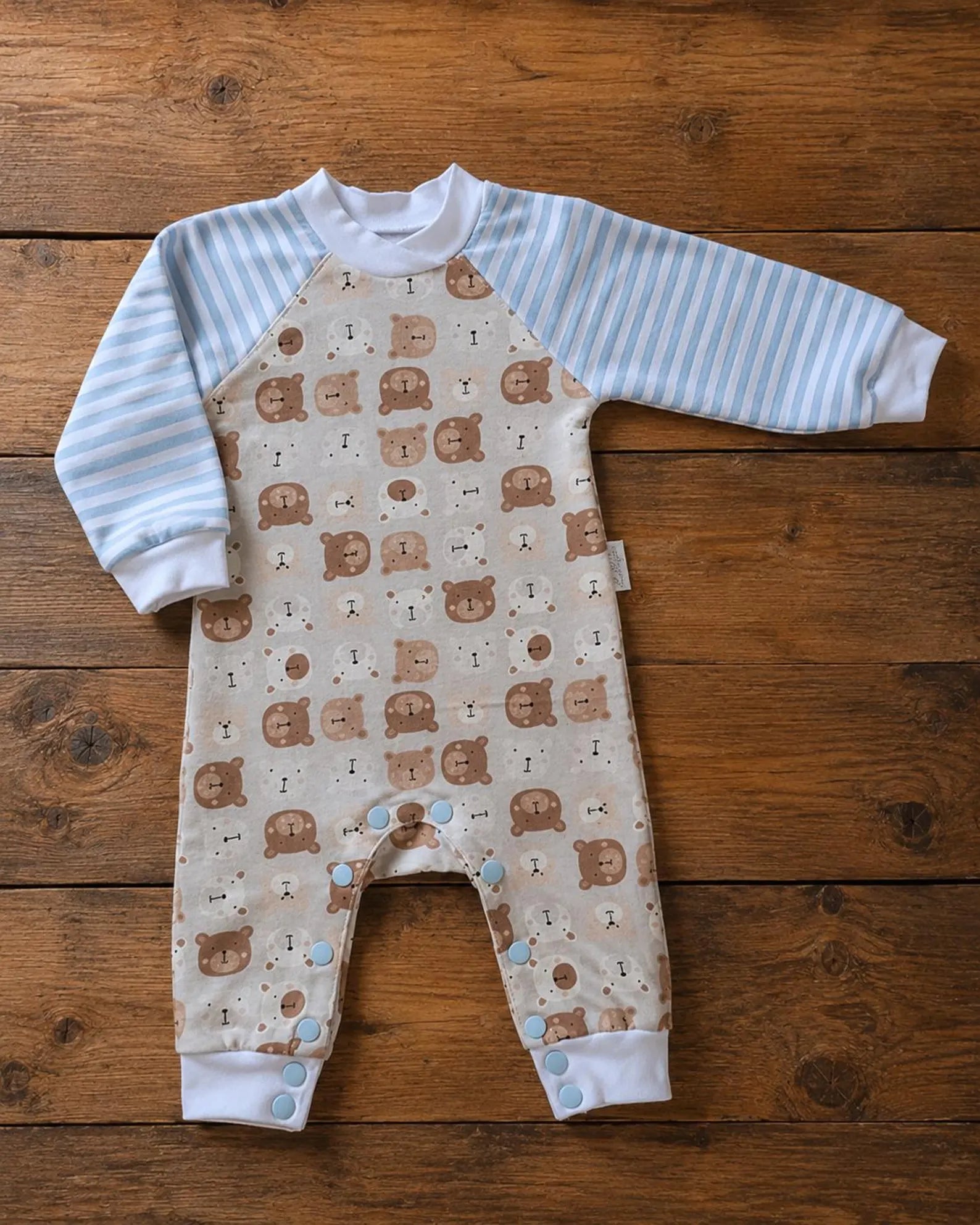 A baby long-sleeve footless romper laid flat on a rustic wooden surface. The romper has light blue and white stripes on the sleeves and collar, a beige body with a brown teddy bear pattern, and white cuffs.