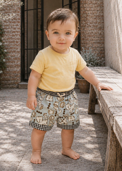 A toddler standing outdoors on a stone patio next to a wooden bench, wearing a pale yellow short-sleeve t-shirt and light blue shorts with a cartoon animal print and black-and-white gingham trim.