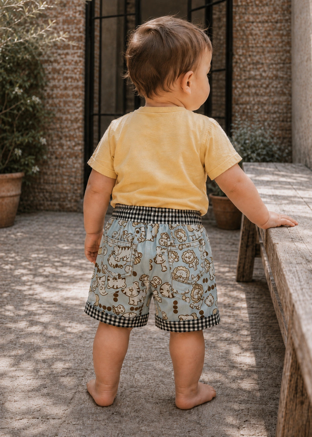 A toddler standing outdoors on a stone patio next to a wooden bench, wearing a pale yellow short-sleeve t-shirt and light blue shorts with a cartoon animal print and black-and-white gingham trim.