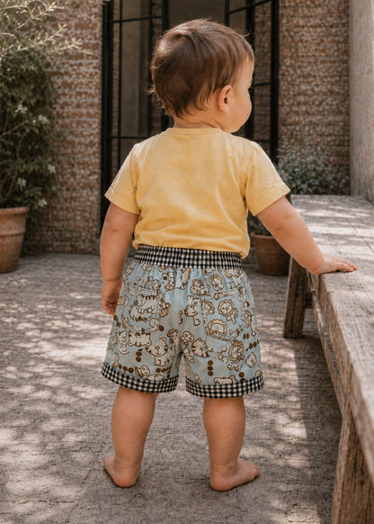 A toddler standing outdoors on a stone patio next to a wooden bench, wearing a pale yellow short-sleeve t-shirt and light blue shorts with a cartoon animal print and black-and-white gingham trim.