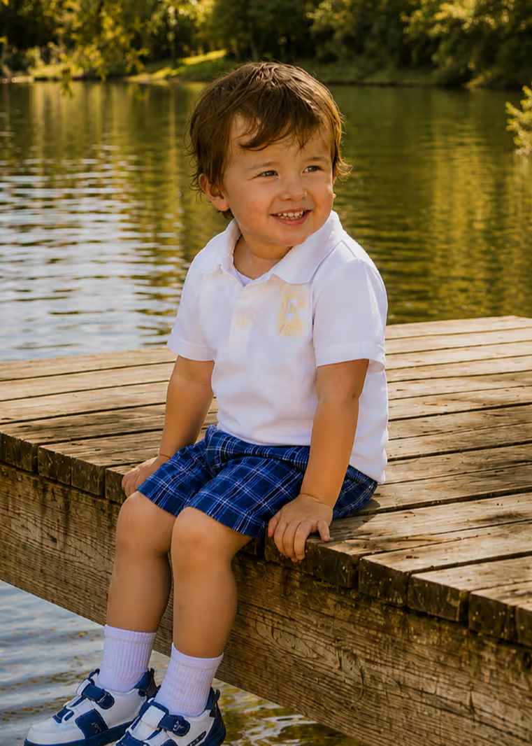 Toddler boy in a white polo shirt and navy plaid Oscar Shorts sitting on a wooden dock by a lake