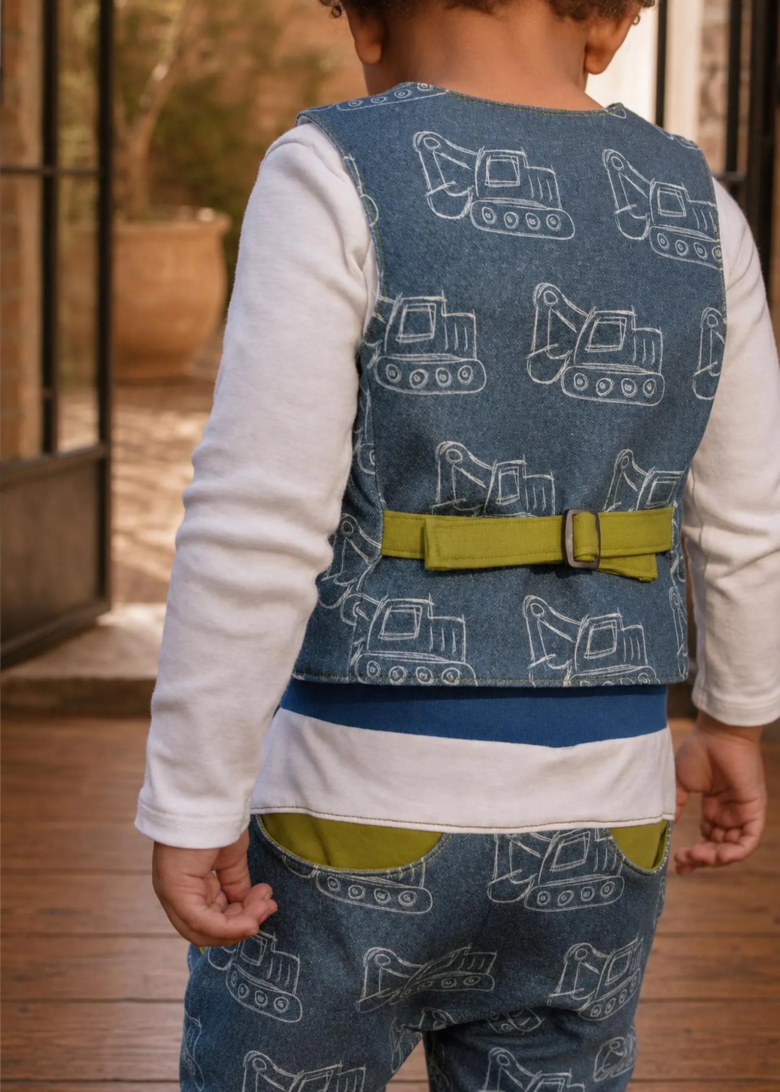 Close-up back view of a toddler wearing a blue excavator print vest featuring a bright green back buckle adjustment.