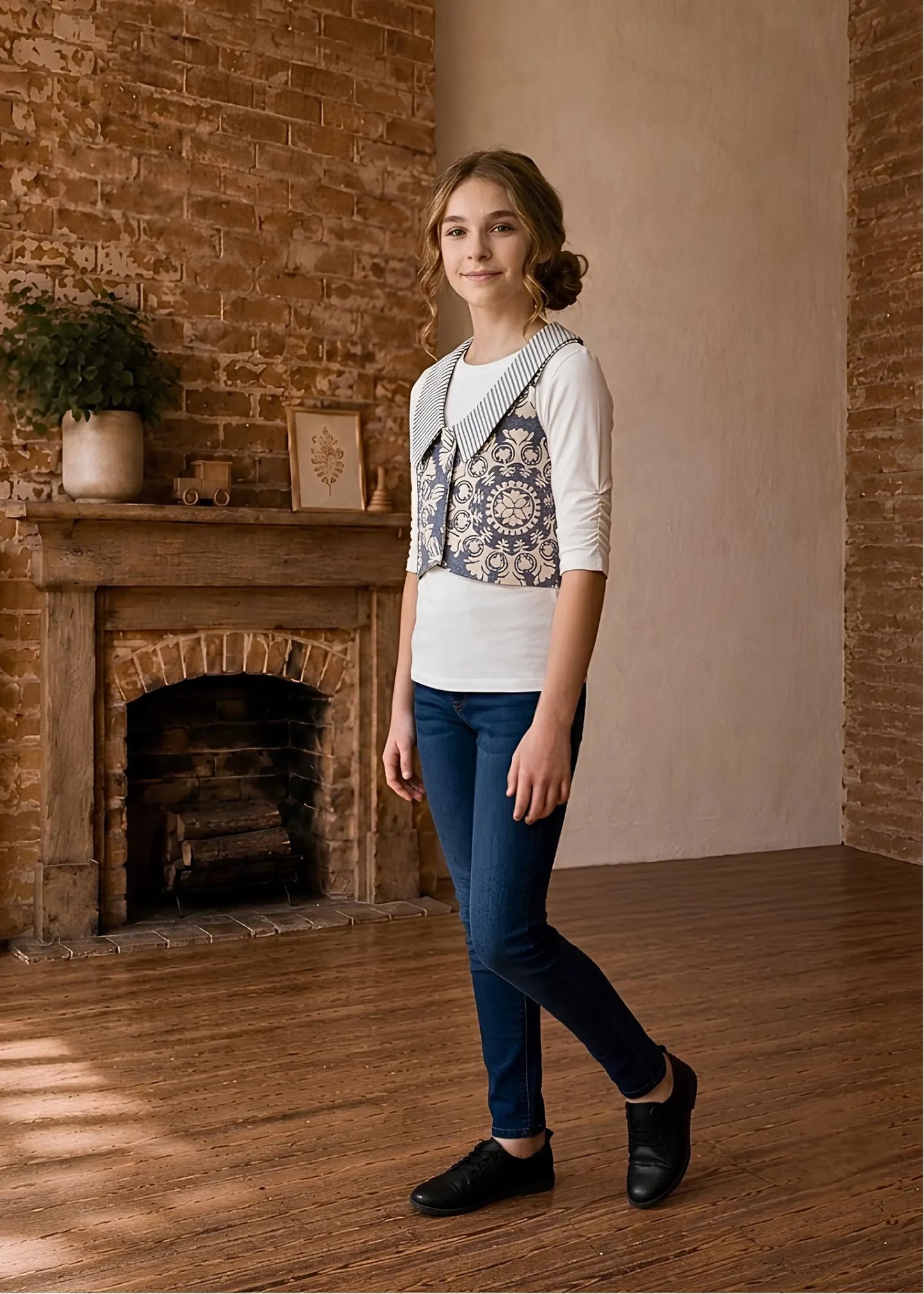 Full-length view of a girl posing in a brick room wearing a printed Brooklyn waistcoat with striped collar over a long sleeve top and jeans.