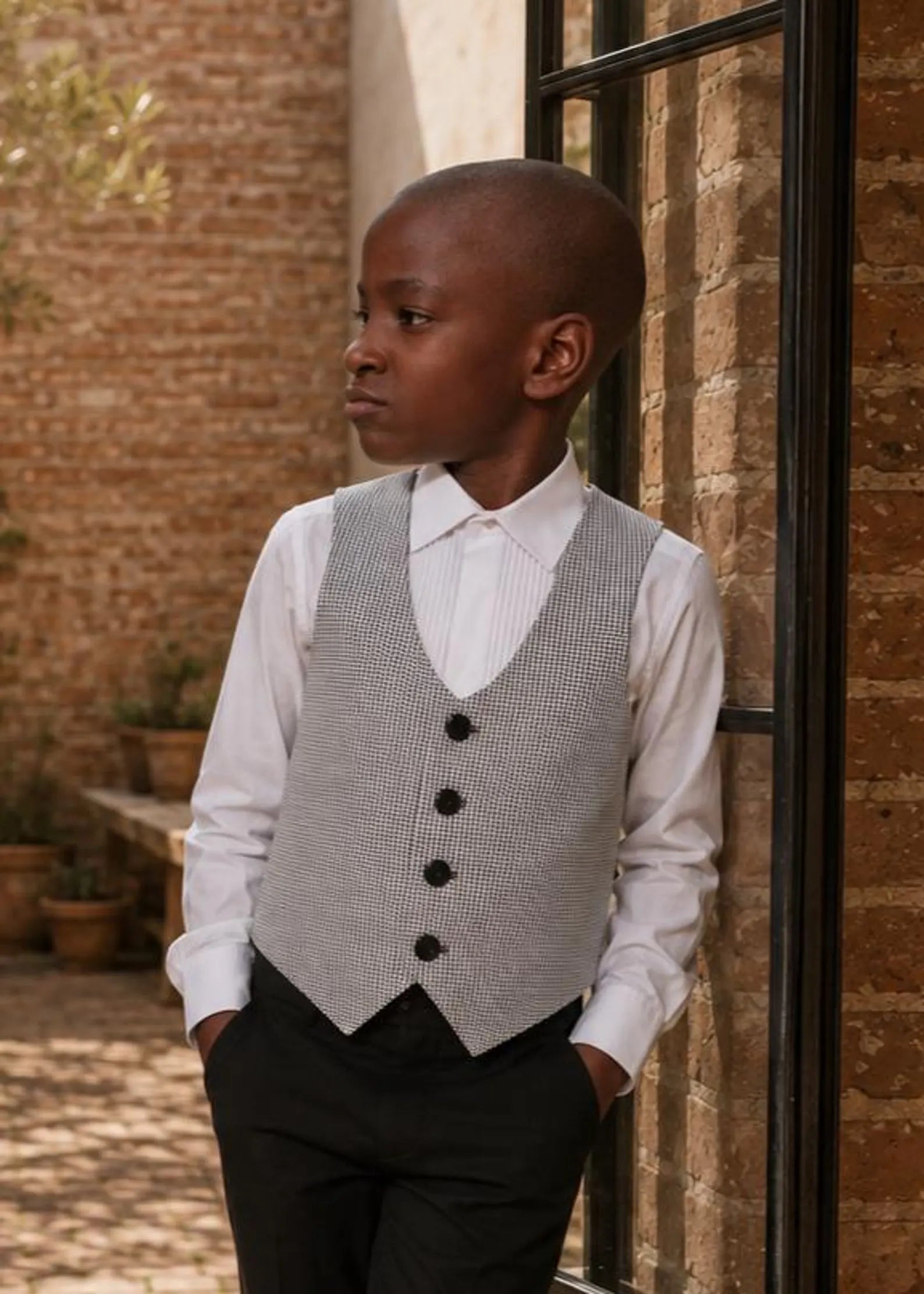 Young boy posing in a formal houndstooth pattern Brooklyn waistcoat with dark buttons over a white dress shirt and black trousers.