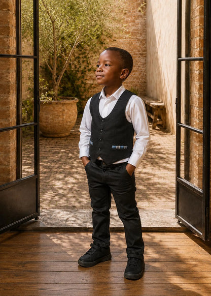 Full-length view of a young boy wearing a navy blue waistcoat with plaid pocket trim, white dress shirt, and black pants standing near a patio doorway.