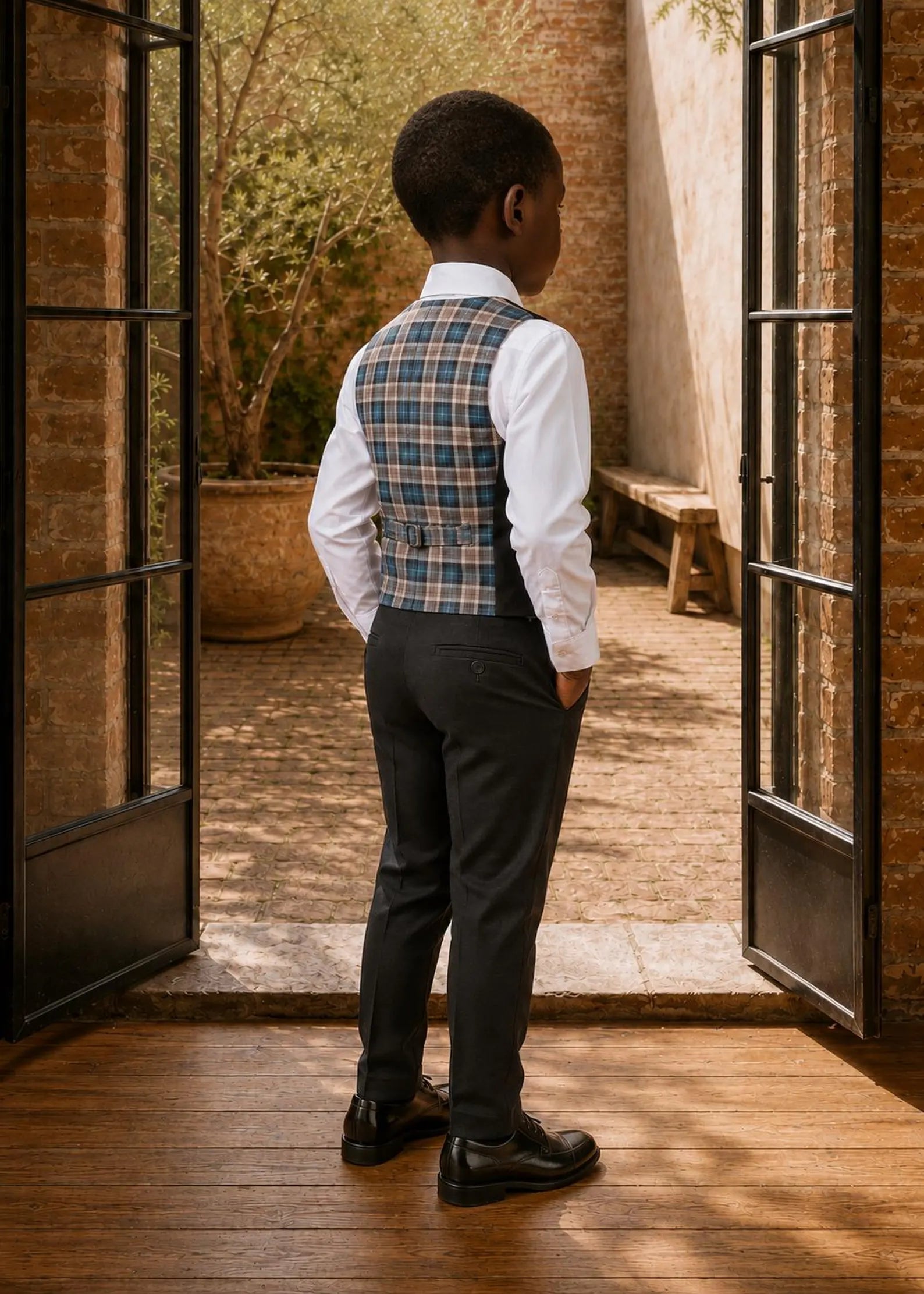 Back view of a boy wearing a plaid pattern Brooklyn waistcoat with an adjustable back buckle strap and dark dress pants.