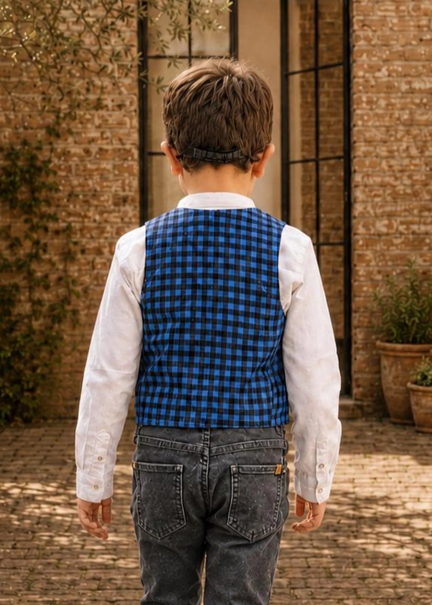 Young boy wearing glasses, a blue and black check Brooklyn waistcoat, white collarless shirt, and black jeans.