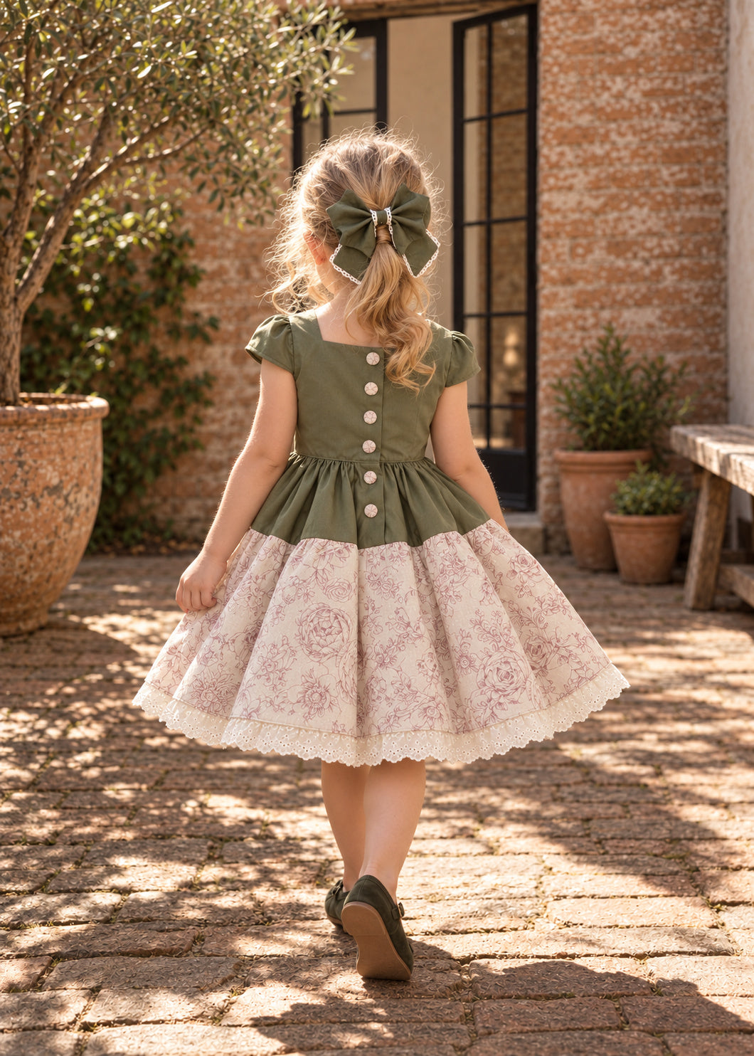 Isabella Dress PDF sewing pattern. Back view of a little girl with a hair bow in a green bodice dress with button back and gathered pink floral skirt with lace hem.