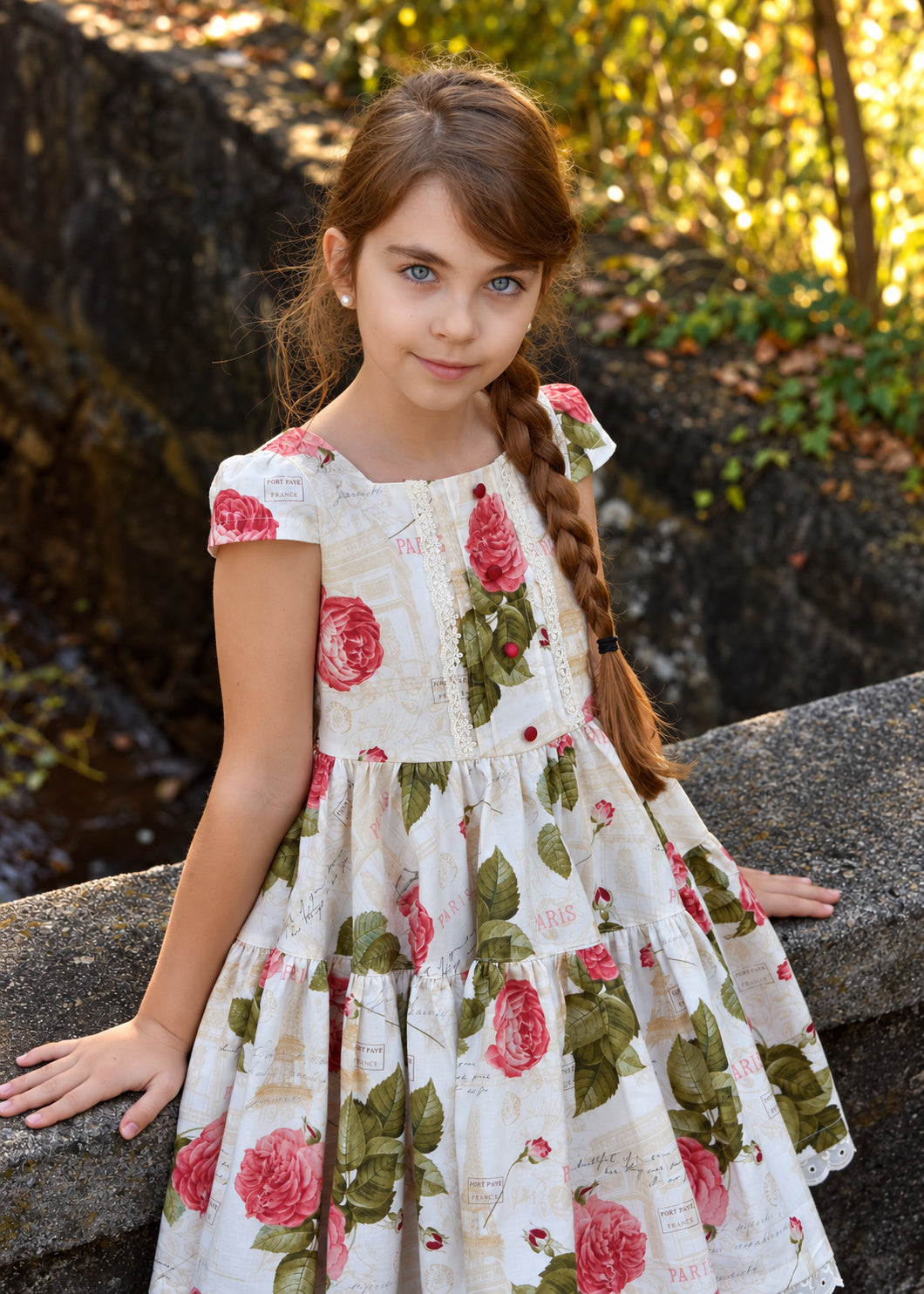 Isabella Dress PDF sewing pattern. Girl with a long braid leaning on a stone wall in the woods, wearing a rose print dress with cap sleeves and square neckline.