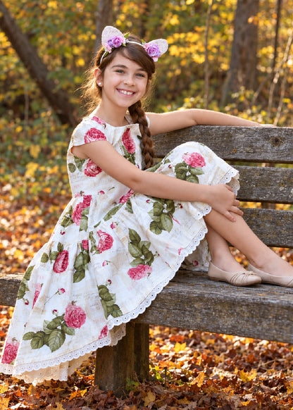Isabella Dress PDF sewing pattern. Smiling girl sitting on a wooden bench among autumn leaves, wearing a rose print dress with lace-trimmed tiered skirt.