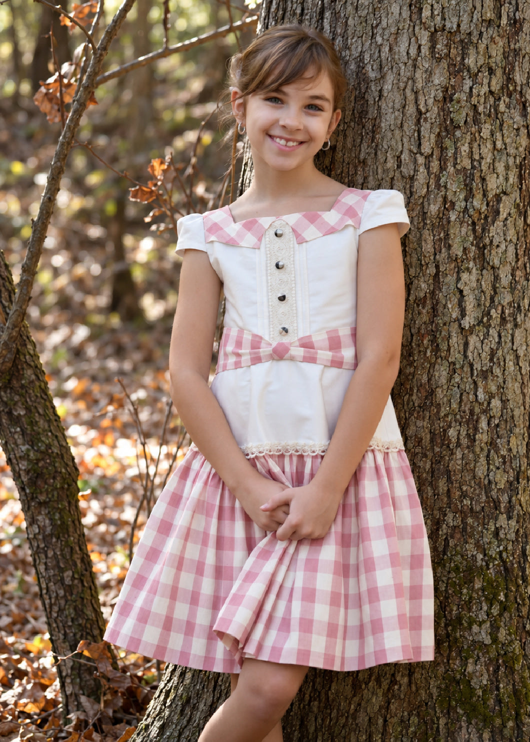 Isabella Dress PDF sewing pattern. Christmas version: smiling girl leaning on a tree in a dress with a white button-front bodice, gingham trim and pink gingham tiered skirt.