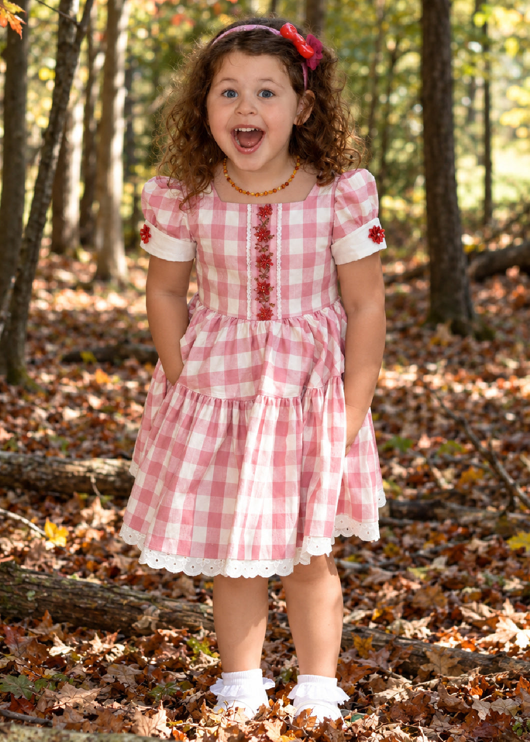 Isabella Dress PDF sewing pattern. Christmas version: laughing curly-haired girl in a pink gingham dress with puff sleeves, red buttons and a lace-trimmed tiered skirt.
