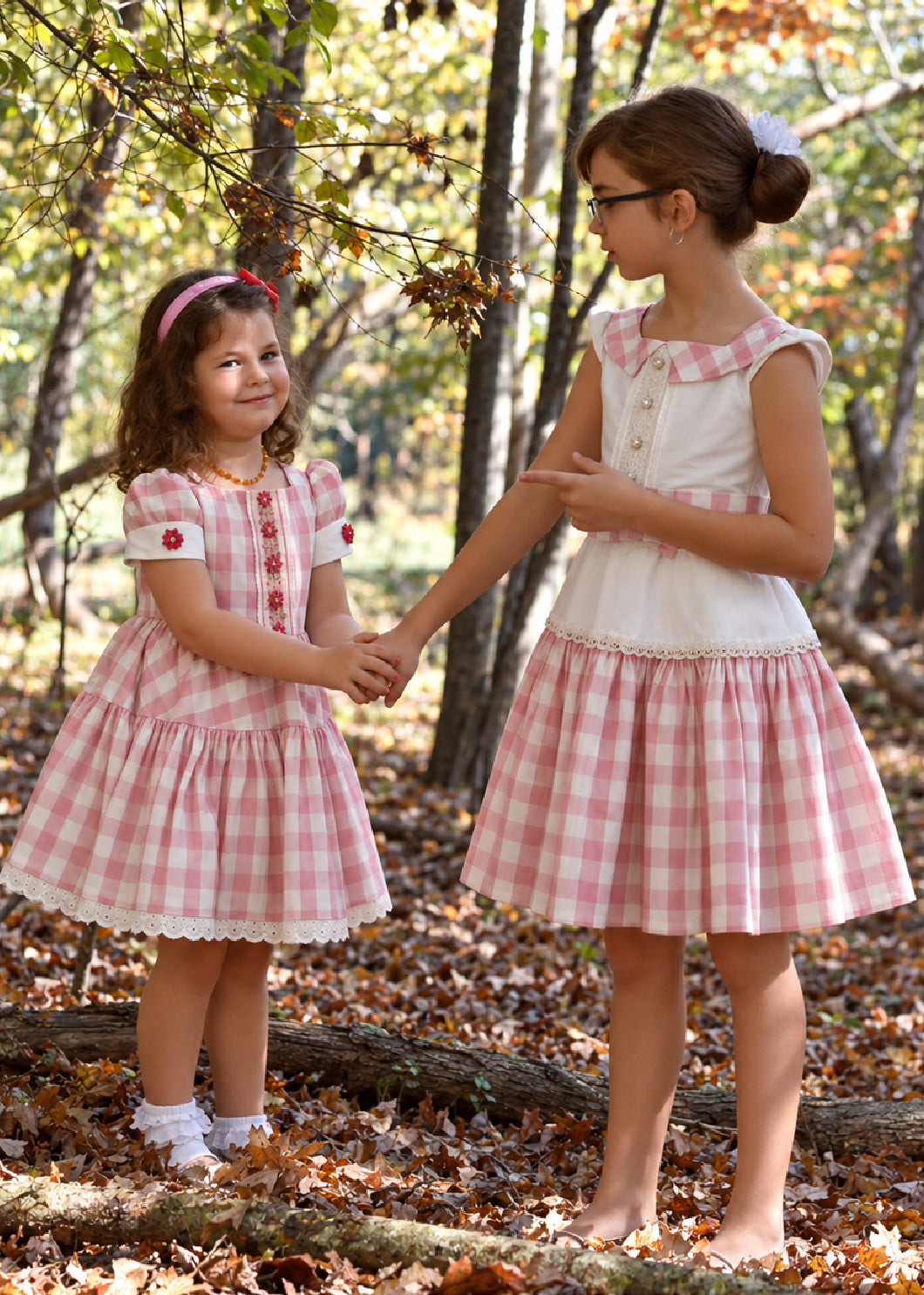 Isabella Dress PDF sewing pattern. Christmas version: two sisters holding hands in the woods wearing matching pink gingham Isabella dresses in different sizes.