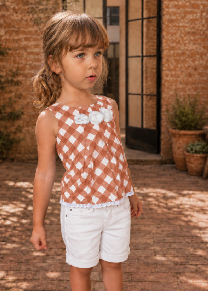 Girl wearing a terracotta check sleeveless top with white flower trim made with the Lily blouse PDF sewing pattern by Frocks & Frolics, paired with white Maisy shorts