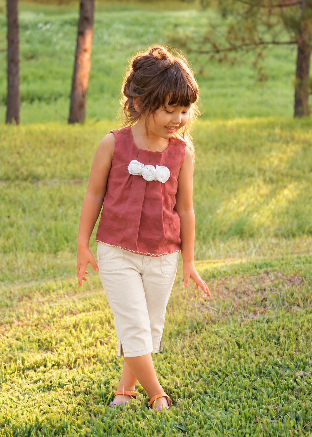 Girl wearing a rust red sleeveless top with front pleats and a white fabric flower, made with the Lily blouse PDF sewing pattern by Frocks & Frolics, paired with cream trousers on a sunny lawn