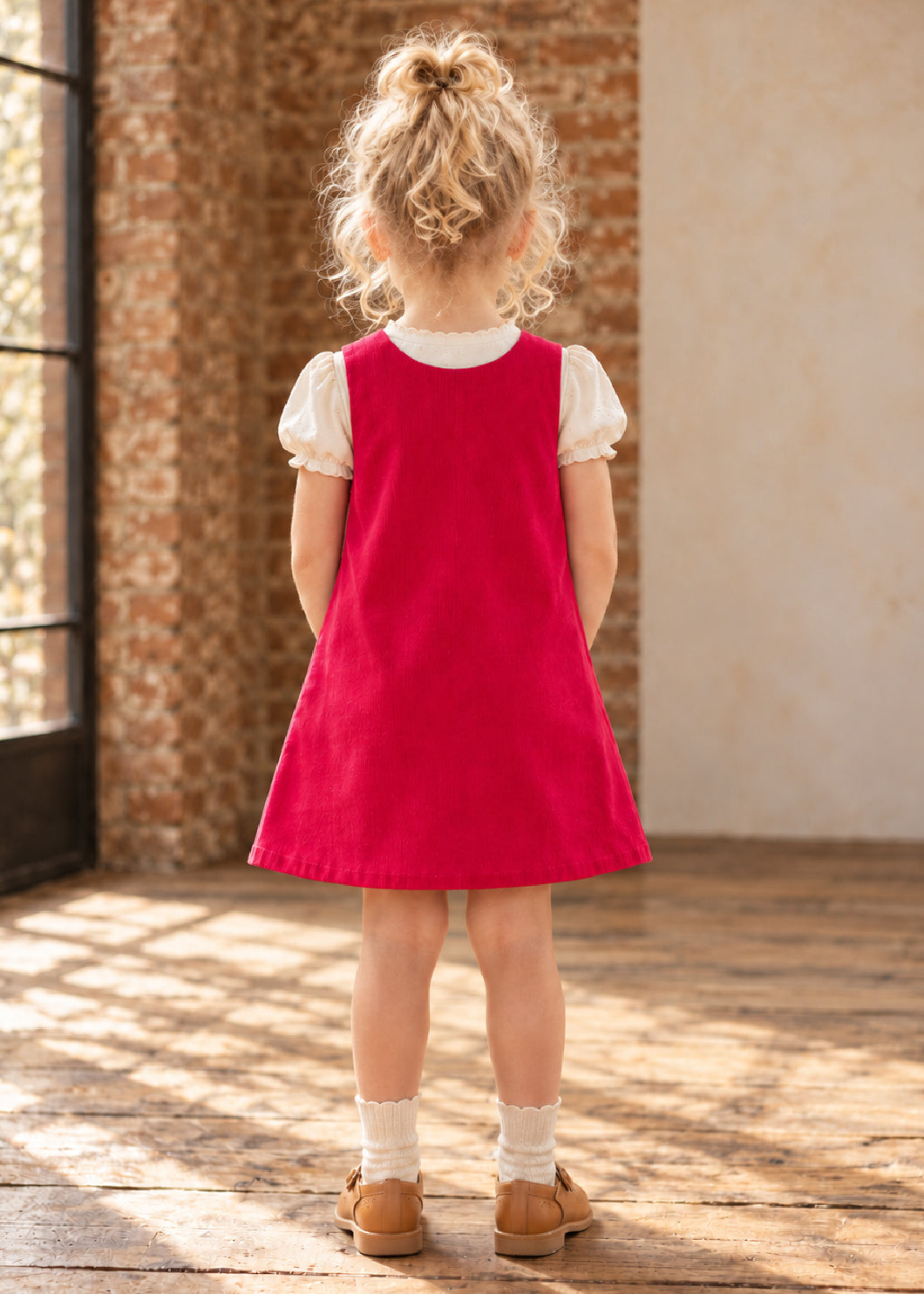 Mimi Pinafore dress PDF sewing pattern. Back view of a girl wearing a red corduroy pinafore dress with button tab closures at the shoulders.