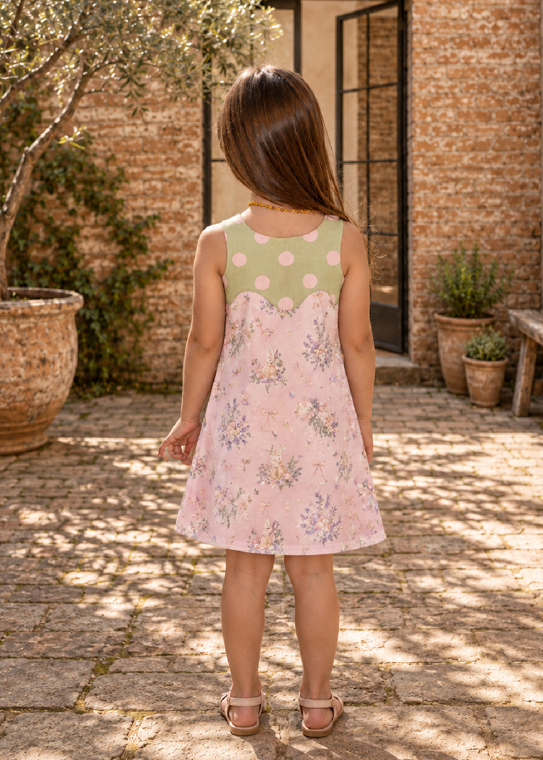 Mimi Pinafore dress PDF sewing pattern. Back view of a girl in a pink floral pinafore dress with a contrast green yoke, standing in a sunny courtyard.