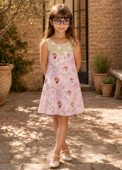 Mimi Pinafore dress PDF sewing pattern. Front view of a girl wearing a pink floral pinafore dress with a contrast green yoke and shoulder button tabs.