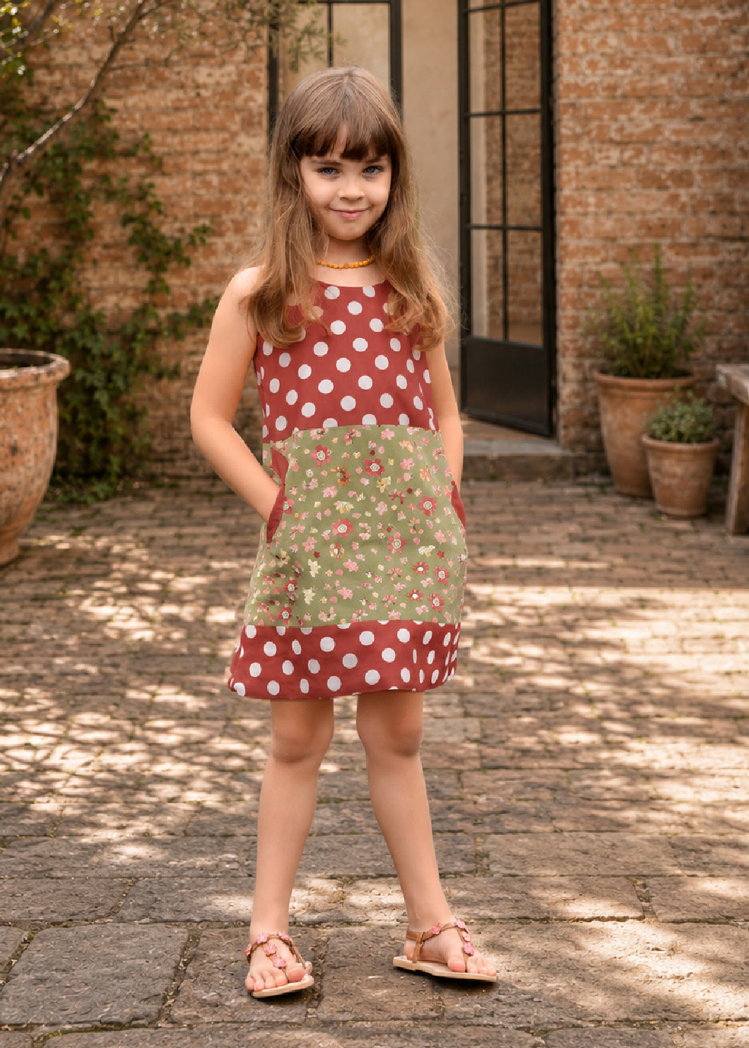 Mimi Pinafore dress PDF sewing pattern. Girl wearing a colour block pinafore with a red polka dot bodice, green floral skirt and round feature pocket.