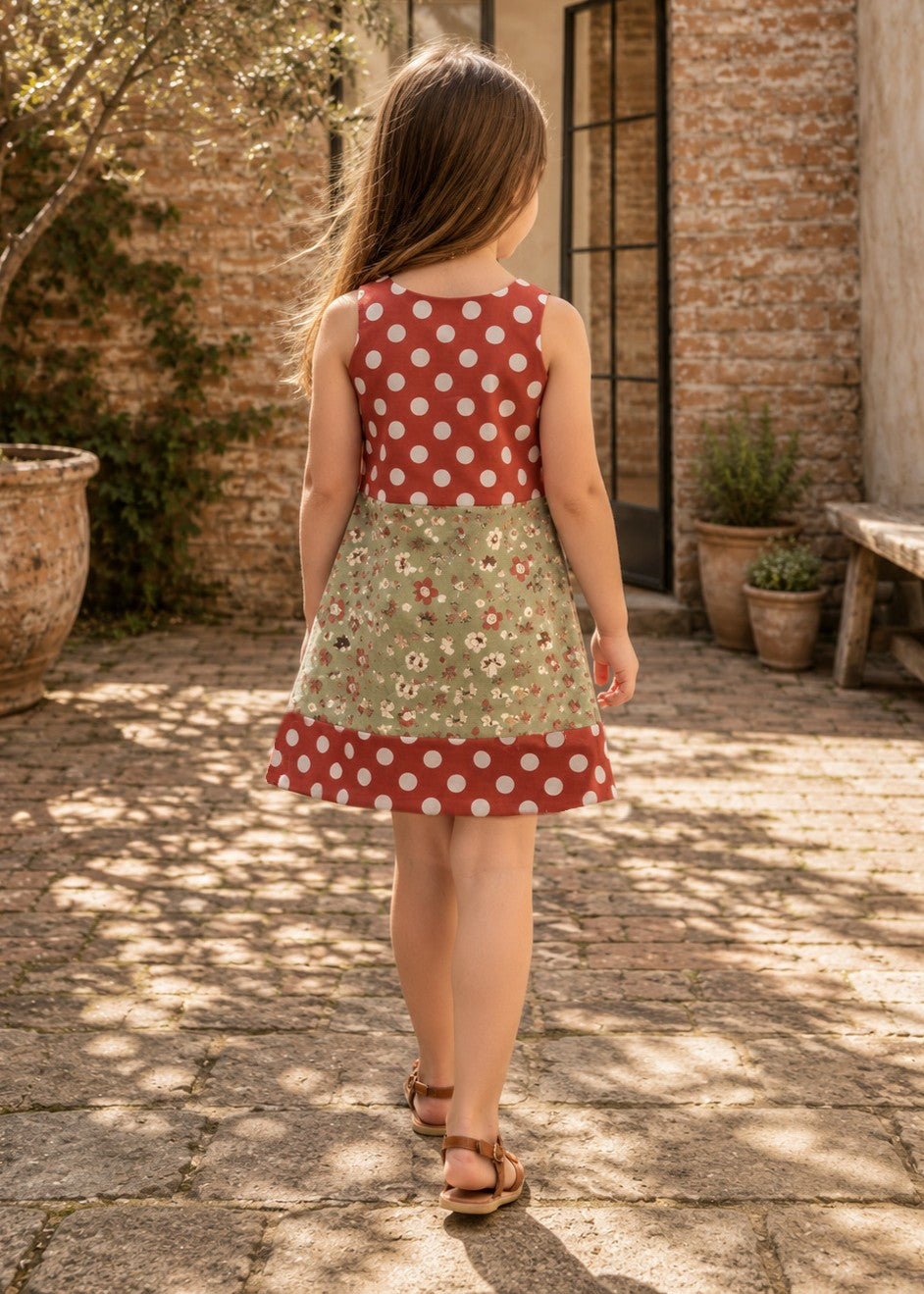 Mimi Pinafore dress PDF sewing pattern. Back view of a girl in a red polka dot and green floral colour block pinafore dress with shoulder tab closures.