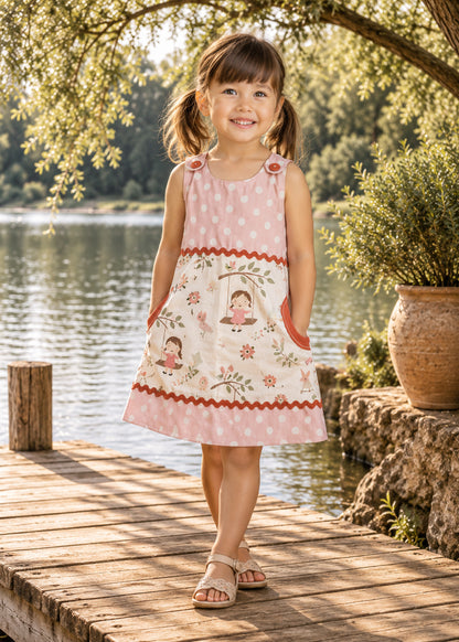 Mimi Pinafore dress PDF sewing pattern. Smiling girl by a lake wearing a pink gingham pinafore with a floral front panel, round pockets and red ric rac trim.