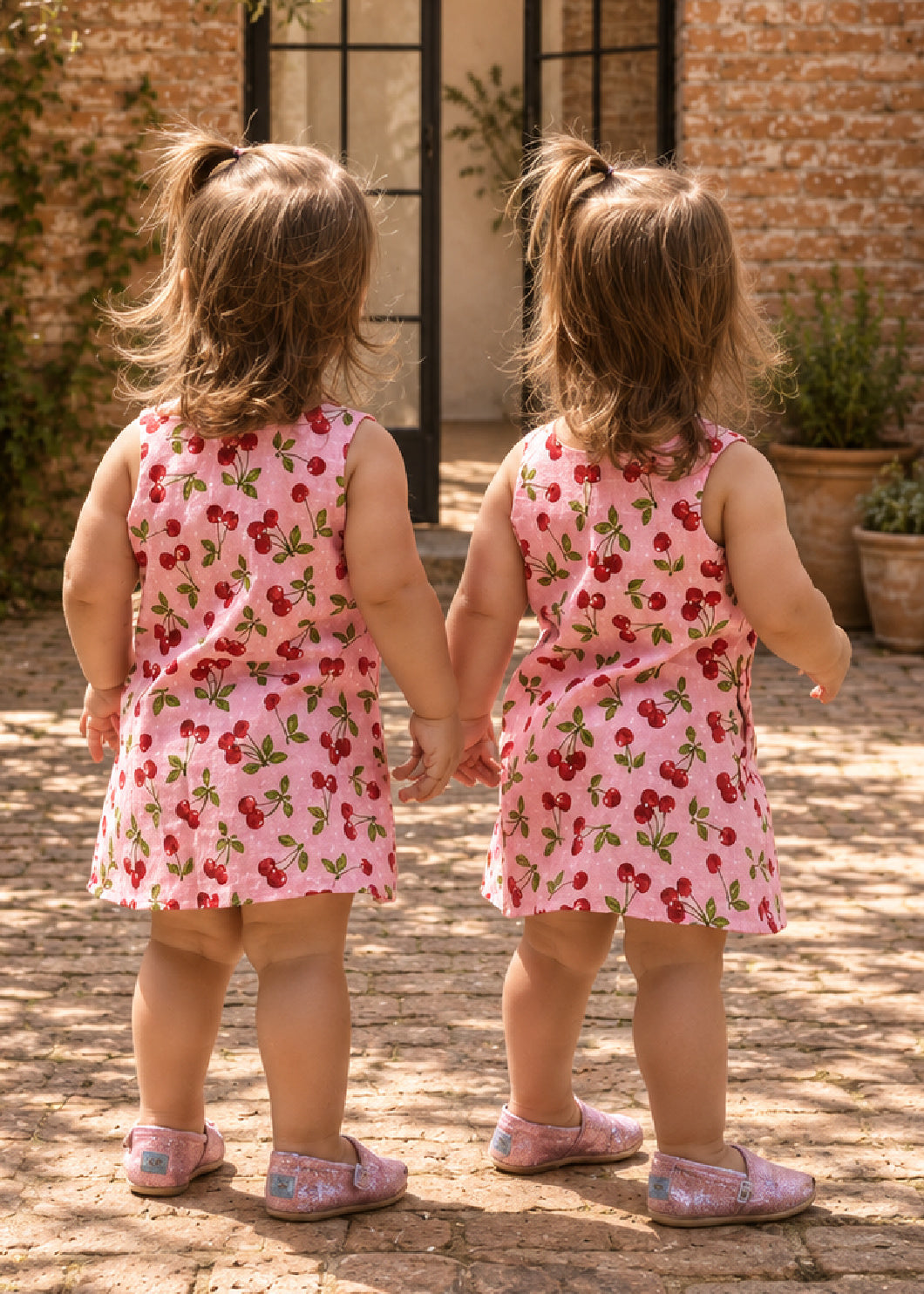 Mimi Pinafore dress PDF sewing pattern. Back view of two toddler girls holding hands in matching pink strawberry print pinafore dresses.