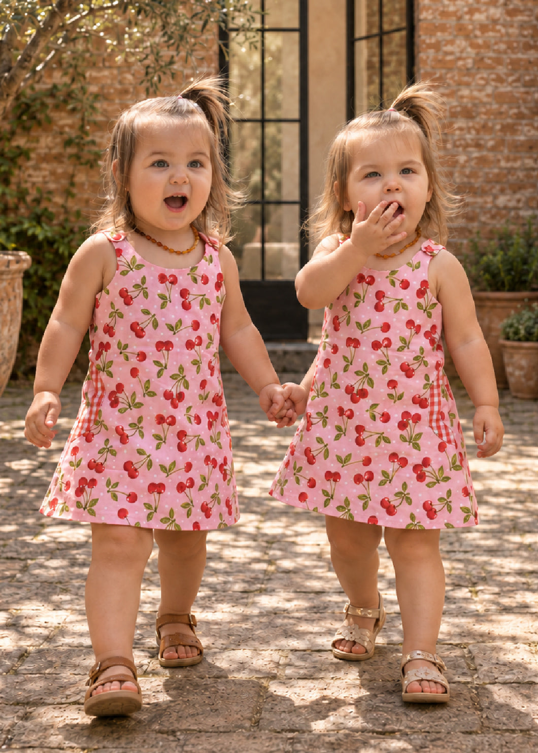 Mimi Pinafore dress PDF sewing pattern. Two toddler girls holding hands in matching pink strawberry print pinafore dresses, front view.