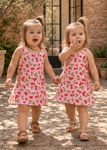 Mimi Pinafore dress PDF sewing pattern. Two toddler girls holding hands in matching pink strawberry print pinafore dresses, front view.