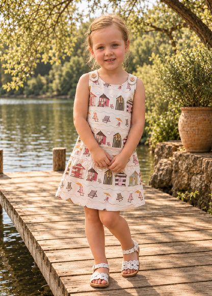 Mimi Pinafore dress PDF sewing pattern. Toddler girl on a lakeside jetty wearing a white pinafore dress with a little house print.