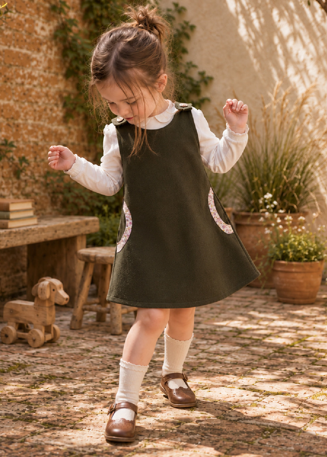 Mimi Pinafore dress PDF sewing pattern. Toddler girl dancing in a dark green pinafore dress with round pockets, worn over a white blouse with a Peter Pan collar.