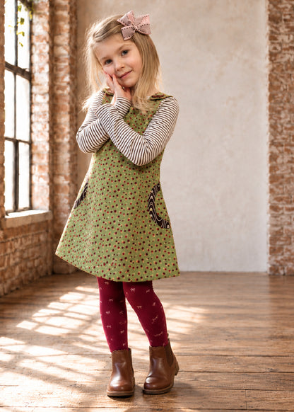 Mimi Pinafore dress PDF sewing pattern. Girl in a green ditsy floral pinafore dress with round pockets, styled with a striped top and red tights.