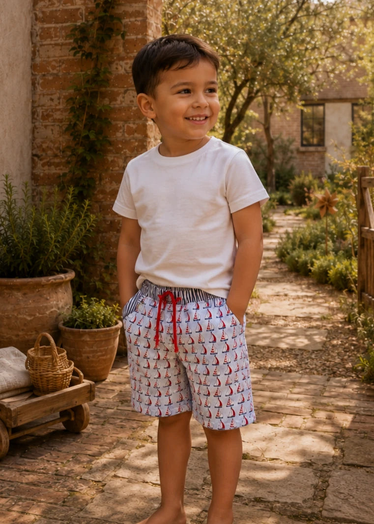 Young boy wearing a white t-shirt and Oscar Shorts in a sailboat-print fabric with a red drawstring, standing on a garden path