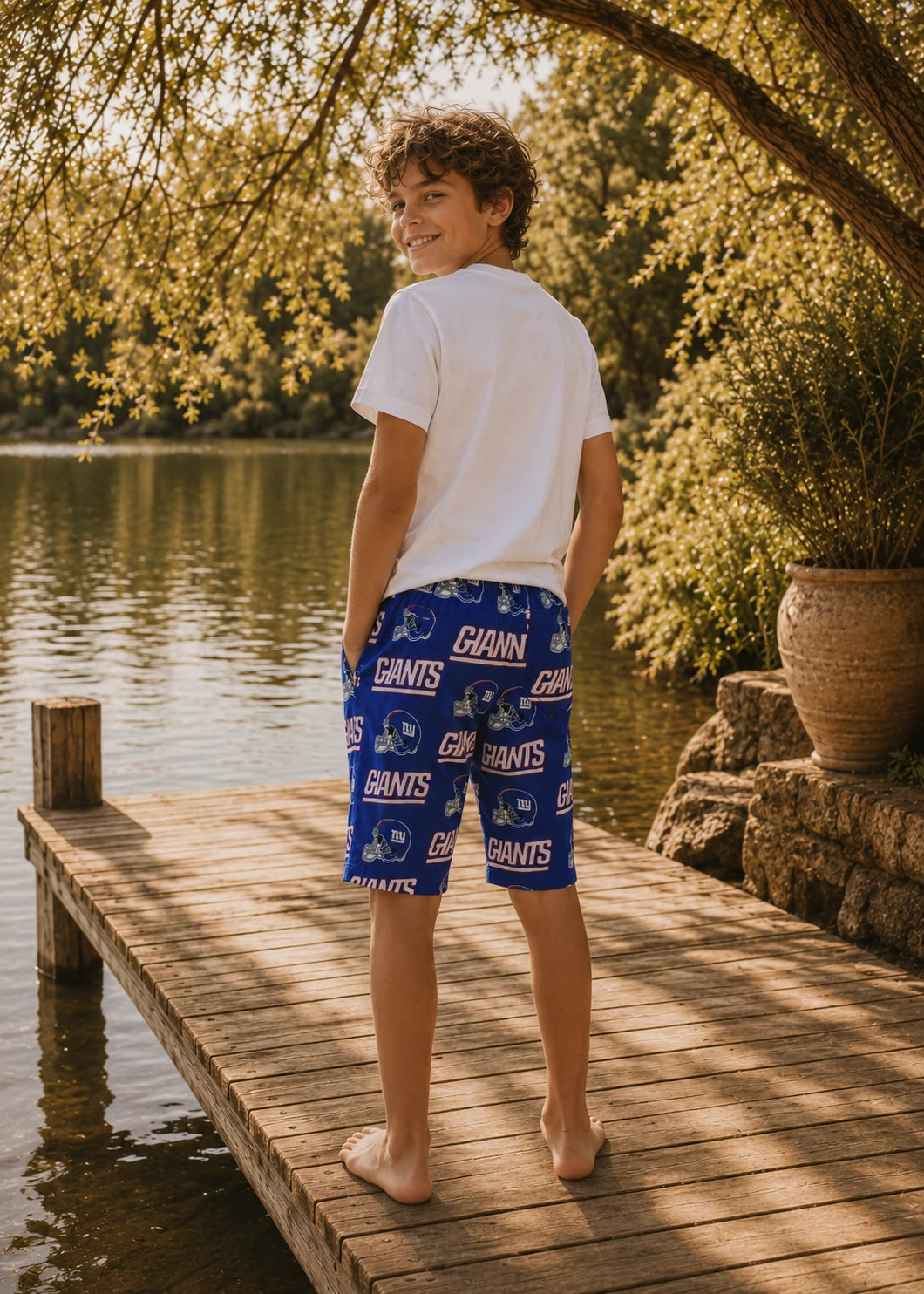 Back view of a preteen boy in a white t-shirt and football team-print Oscar Shorts standing barefoot on a wooden dock