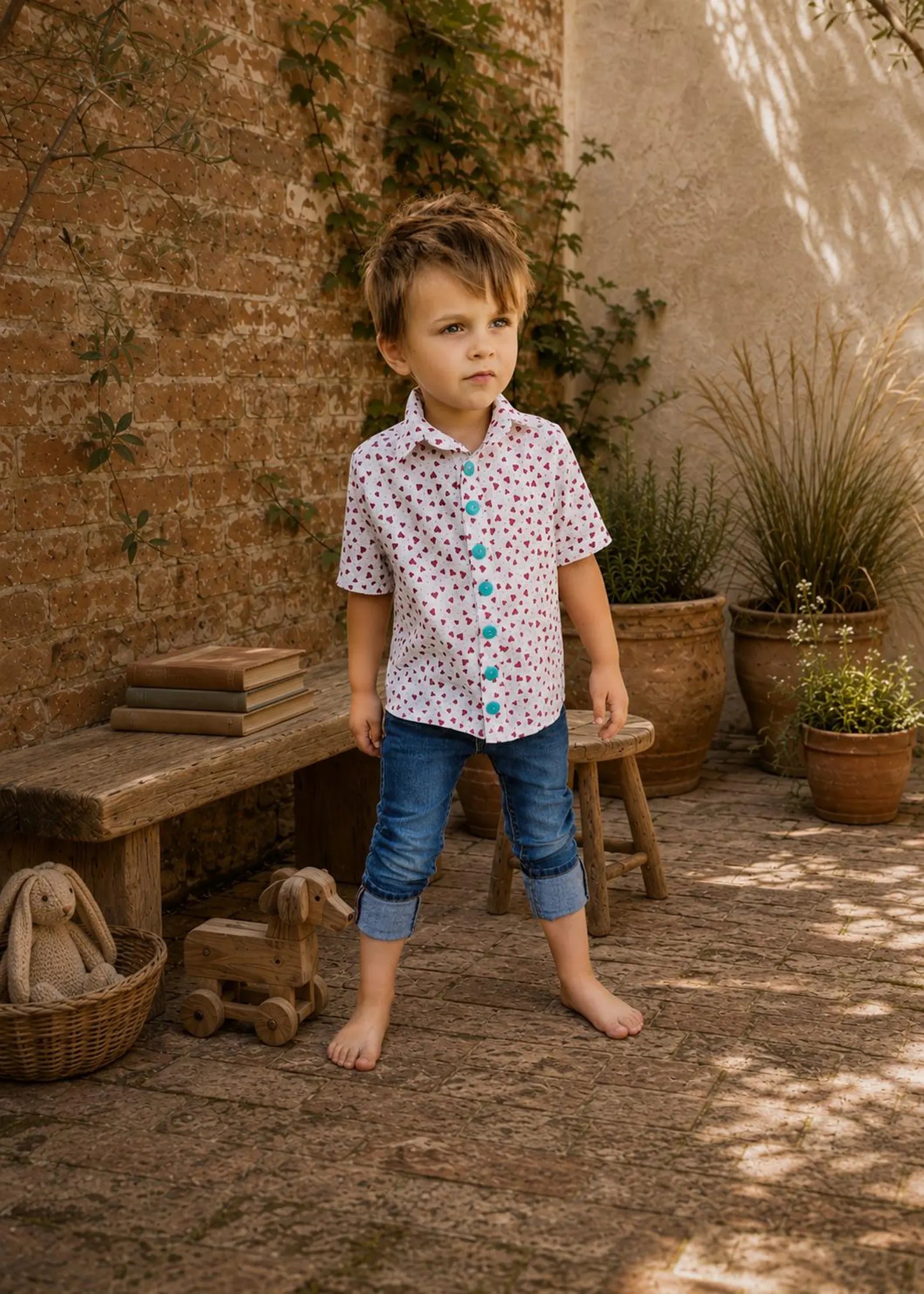 Boy in a red heart-patterned button-up shirt and cuffed jeans standing barefoot in a courtyard.